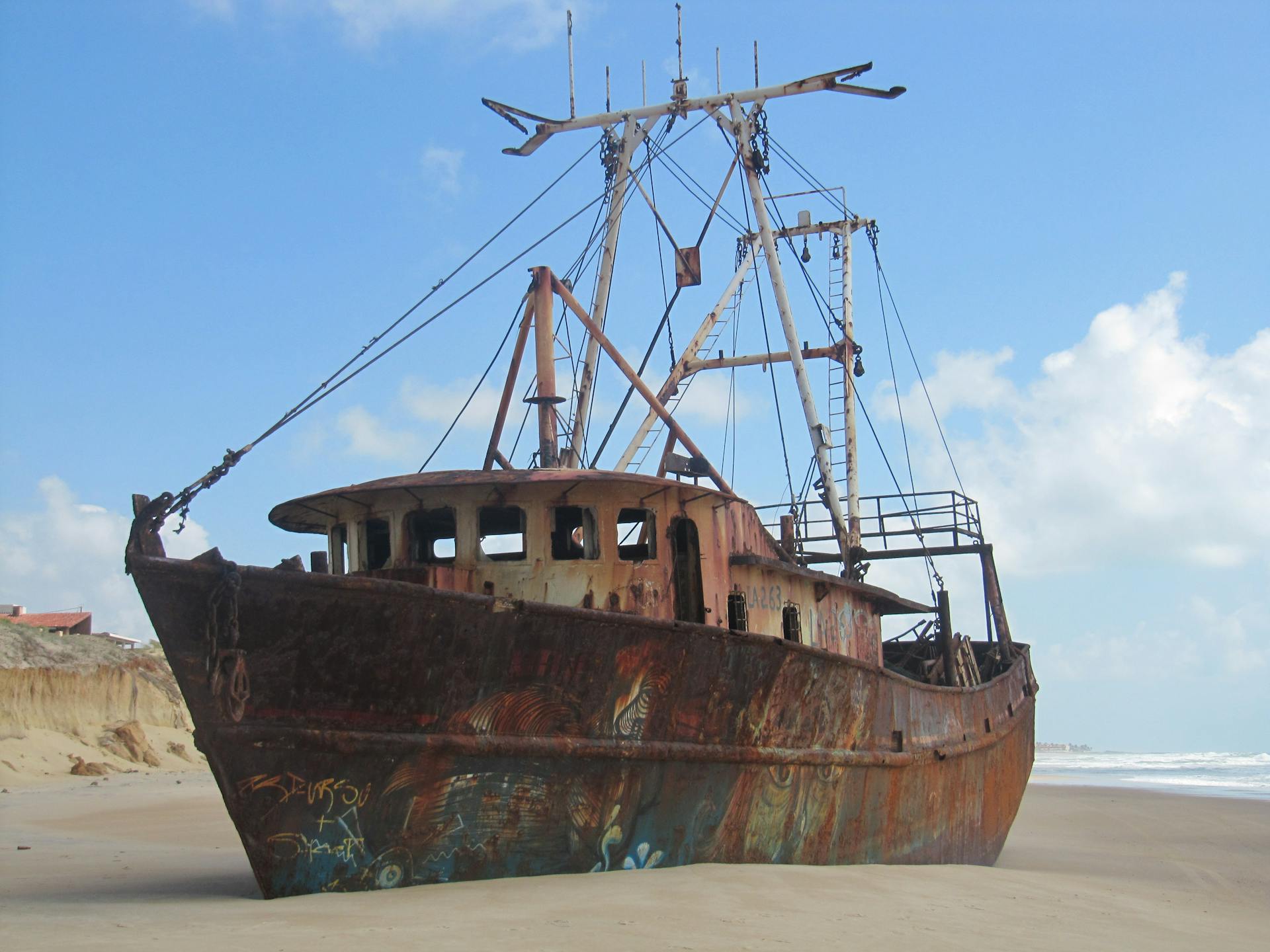 A rusting abandoned shipwreck rests on a sandy beach under a bright blue sky.