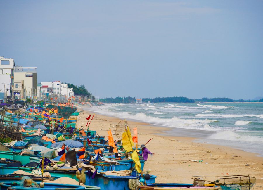 Vibrant fishing boats lined up on a sandy beach beside gentle ocean waves.