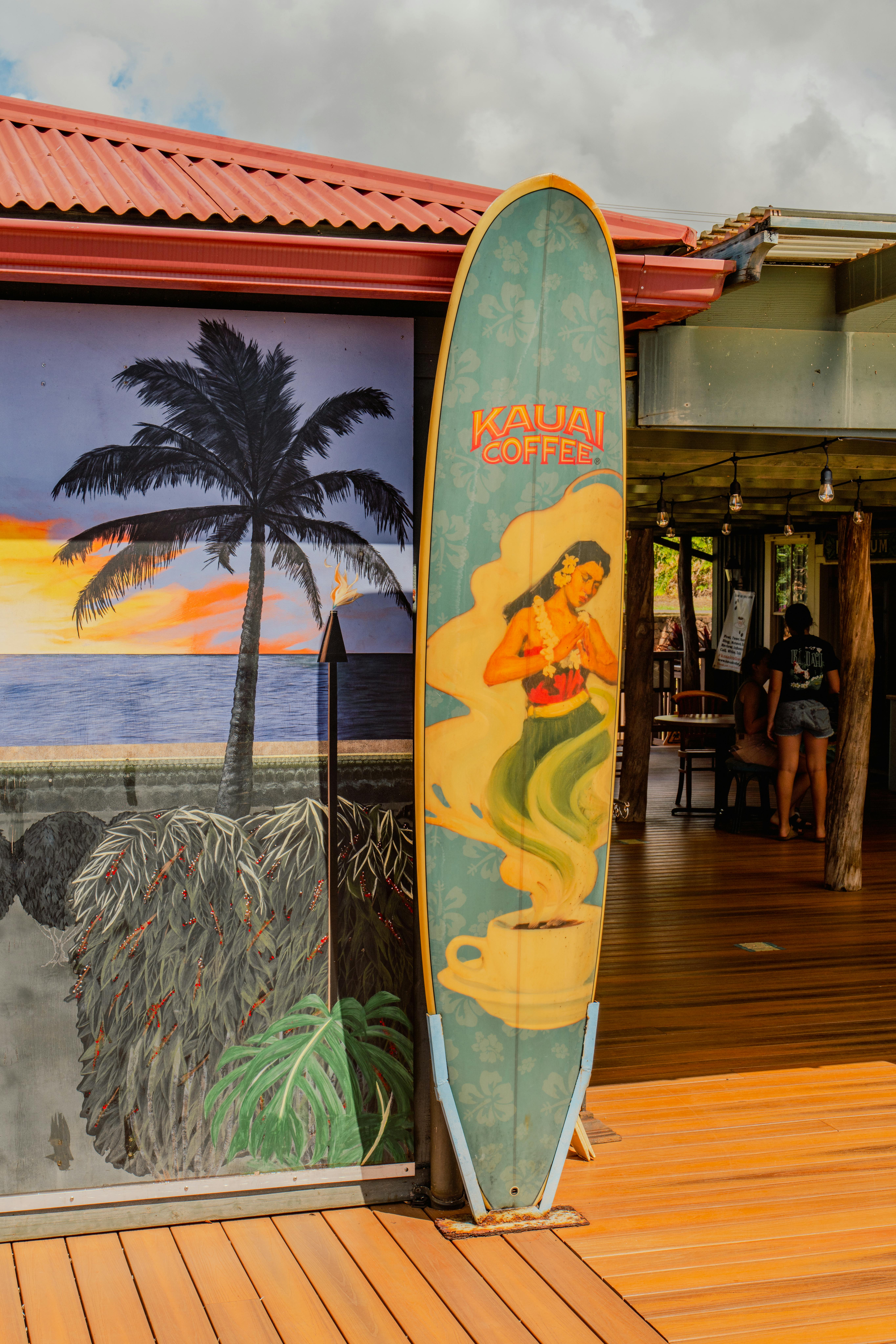 Colorful surfboard with Kauai Coffee branding at a Hawaiian café with a mural backdrop.