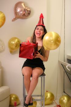 Cheerful woman in party hat holding a gift among golden balloons. Indoor celebration setting.