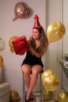 Smiling woman celebrates birthday with party hat, balloons, and gift indoors.