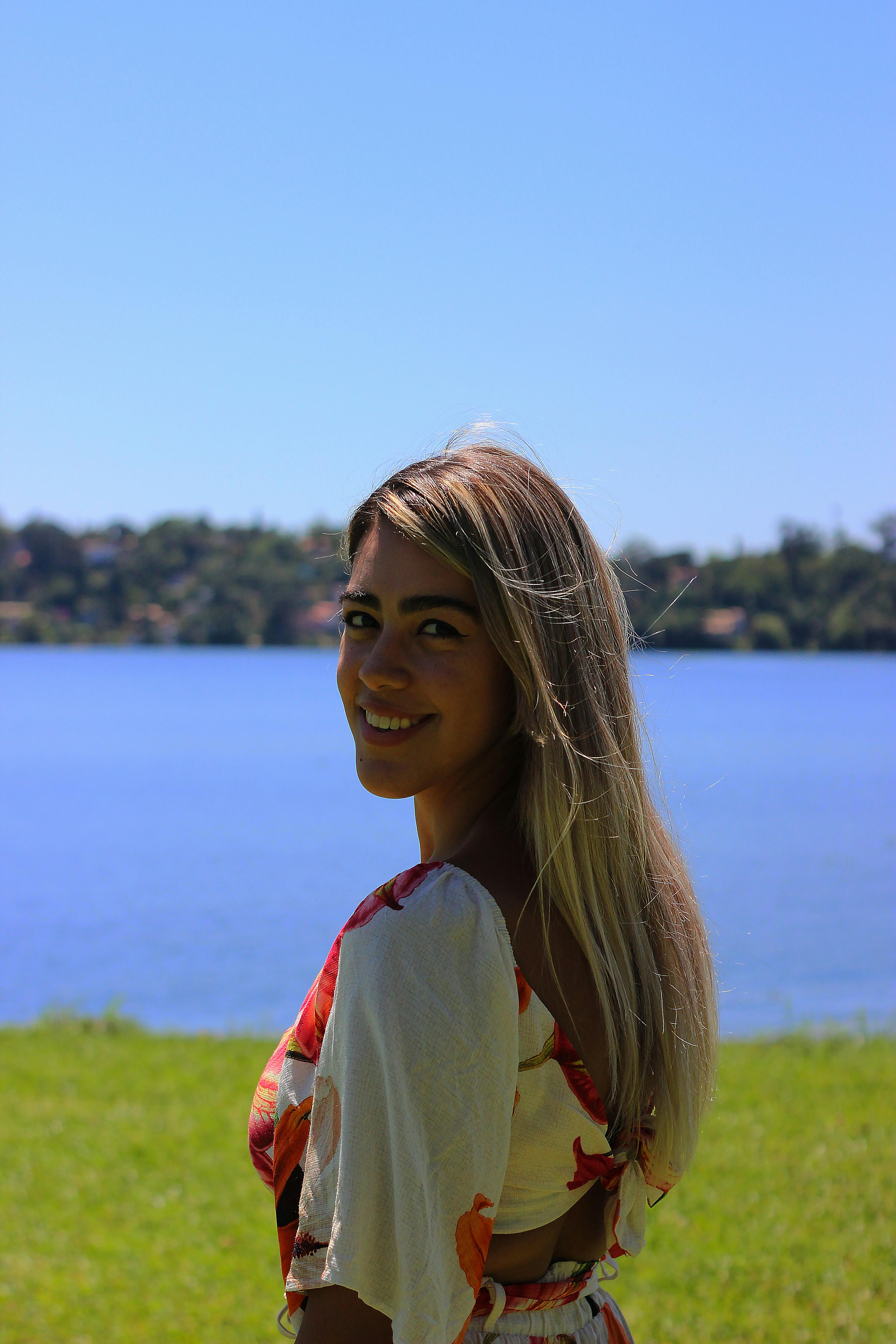 Portrait of a young woman with blond hair smiling by a lake during a sunny day outdoors.