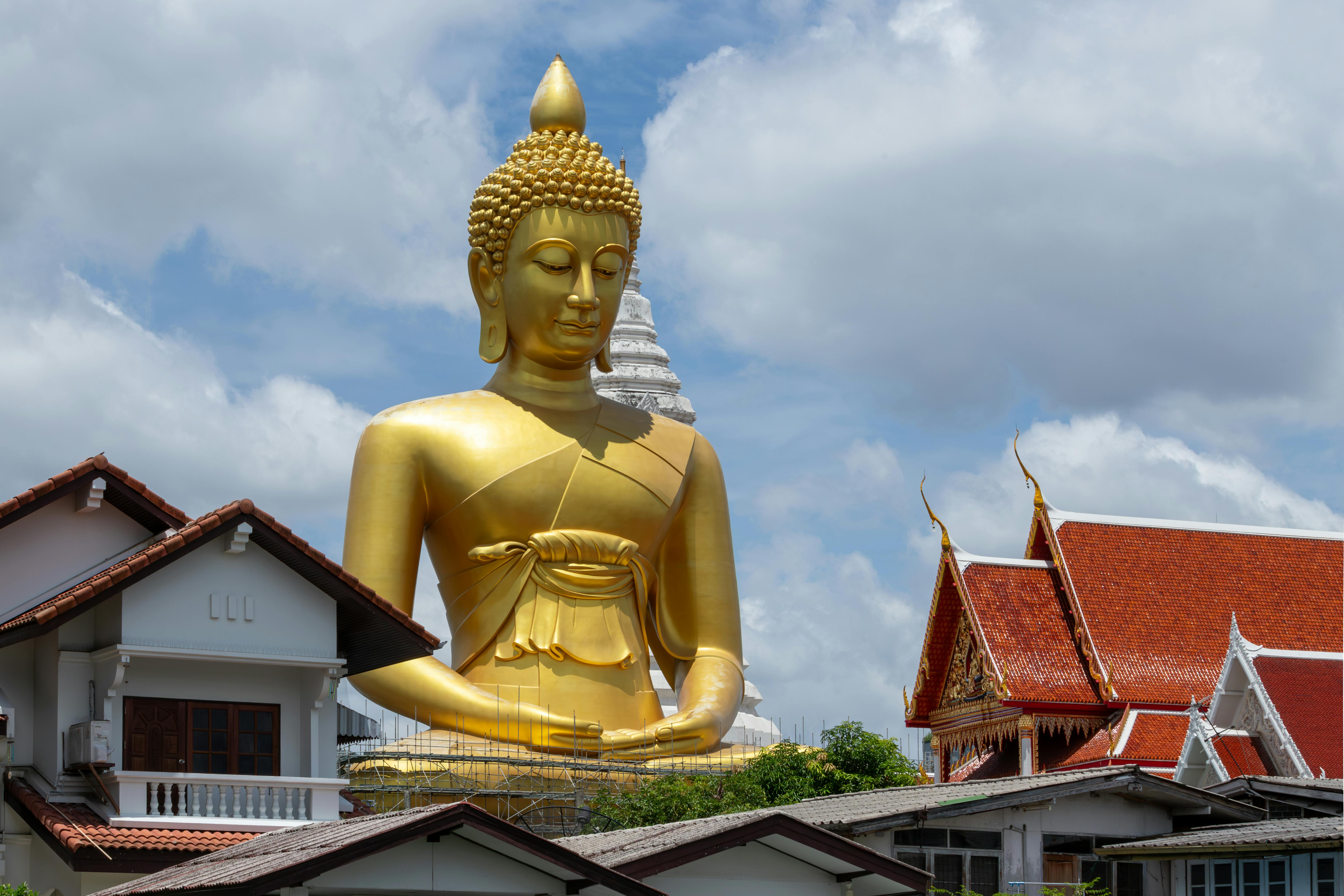 Golden Buddha statue towering over traditional Thai temple with cloudy sky.