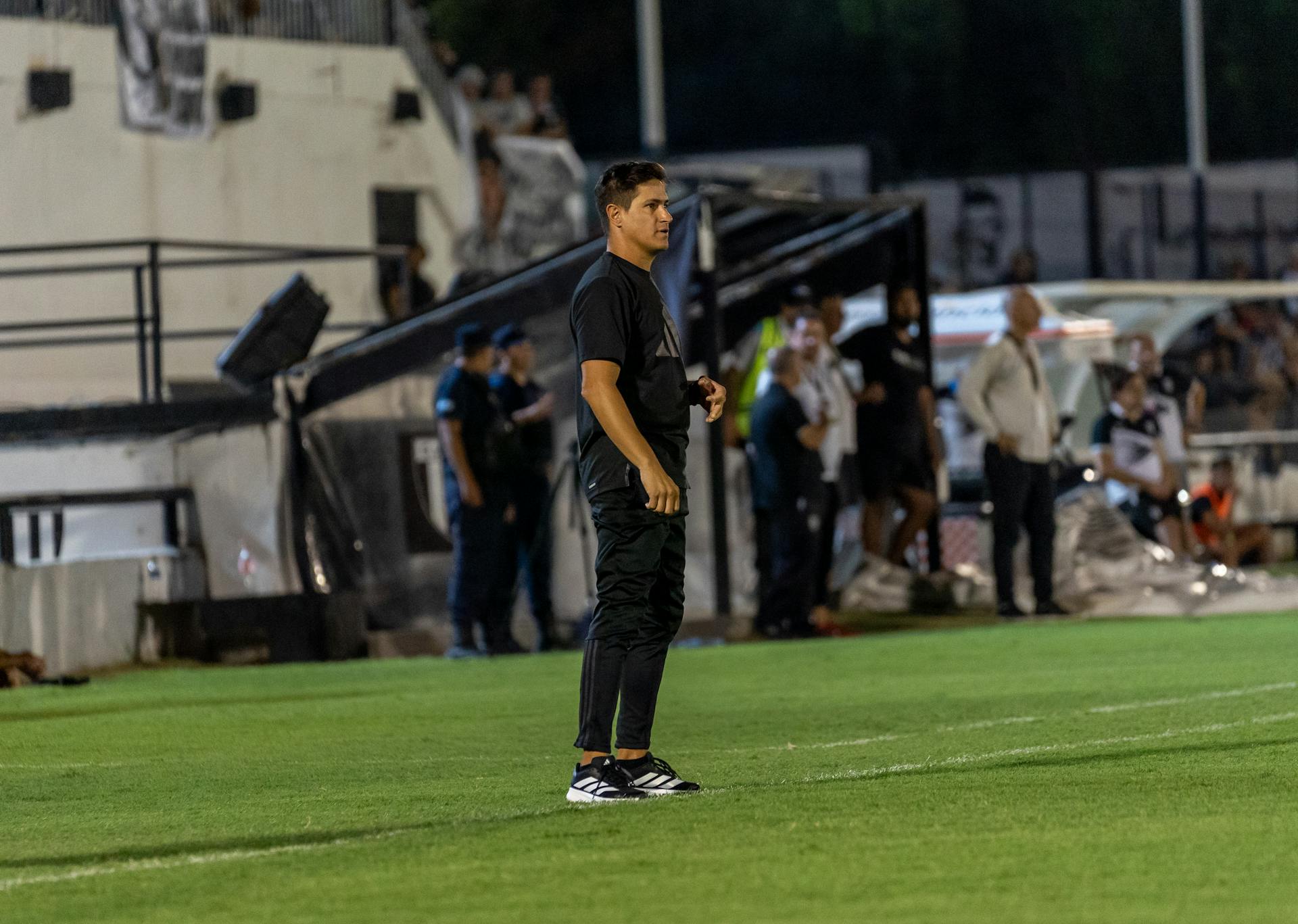 Focused football coach stands on the field sidelines during an intense match at night.