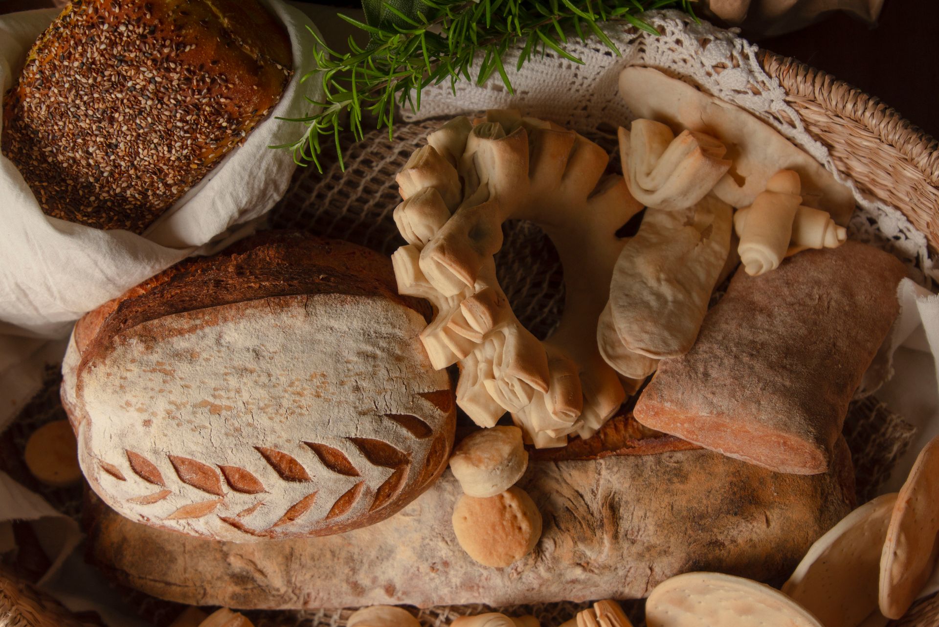 Variety of artisanal breads and pastries arranged on a rustic woven basket with rosemary garnish.
