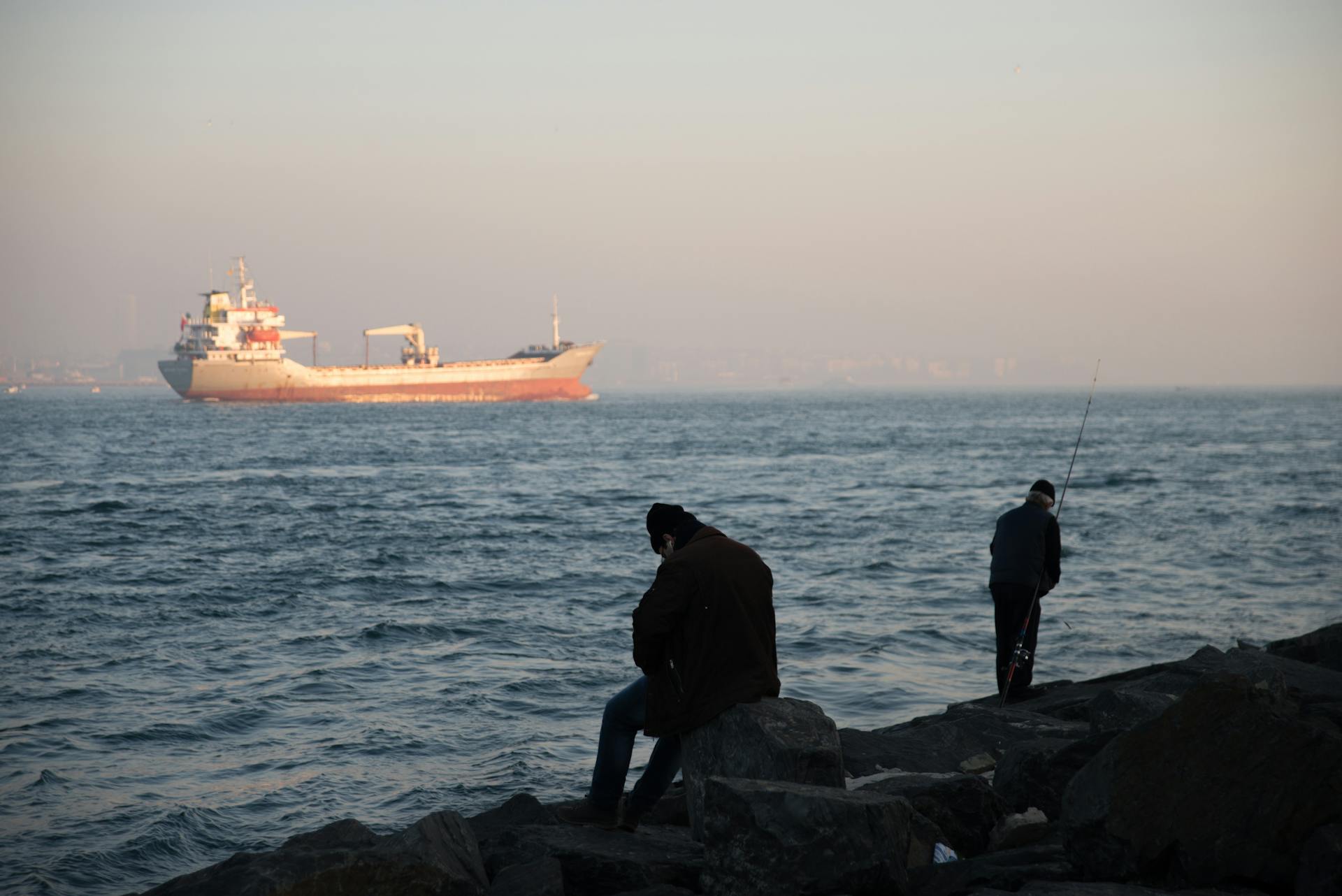 Two men fishing on rocky shore as a cargo ship passes by at dusk.