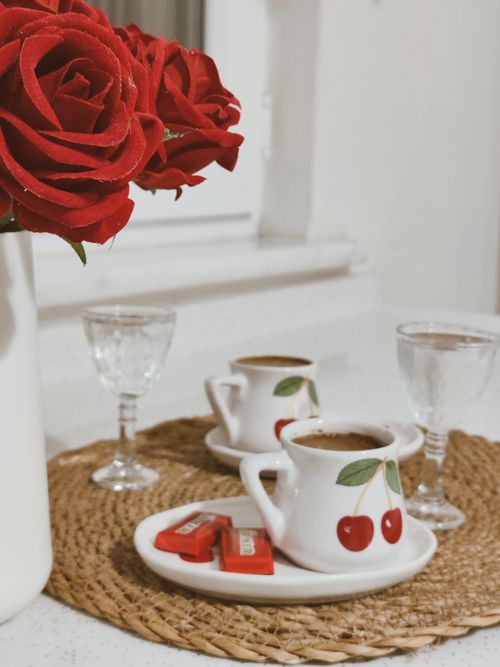 Free A cozy coffee setup featuring red roses and cherry-decorated cups on a woven mat. Stock Photo