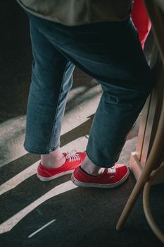 A person stands indoors wearing red sneakers and jeans, sunlight creating shadows on the floor.