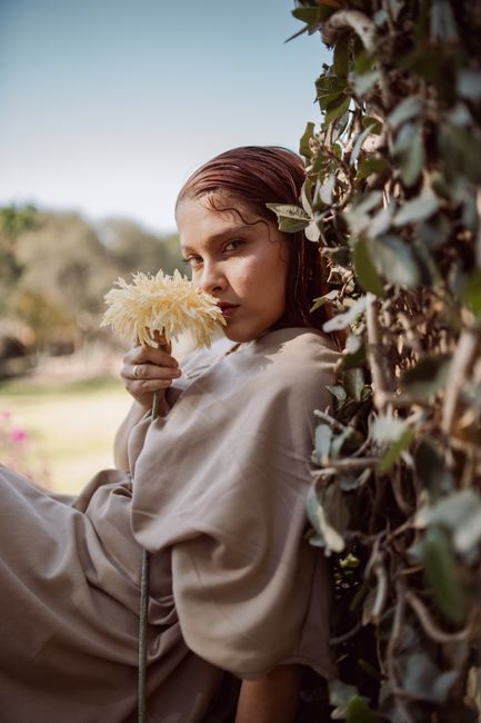 A serene portrait of a woman holding a flower by lush greenery outdoors.