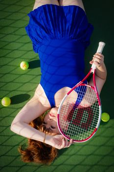 Woman in a blue dress lying on a tennis court with a racket, evoking style and sport.