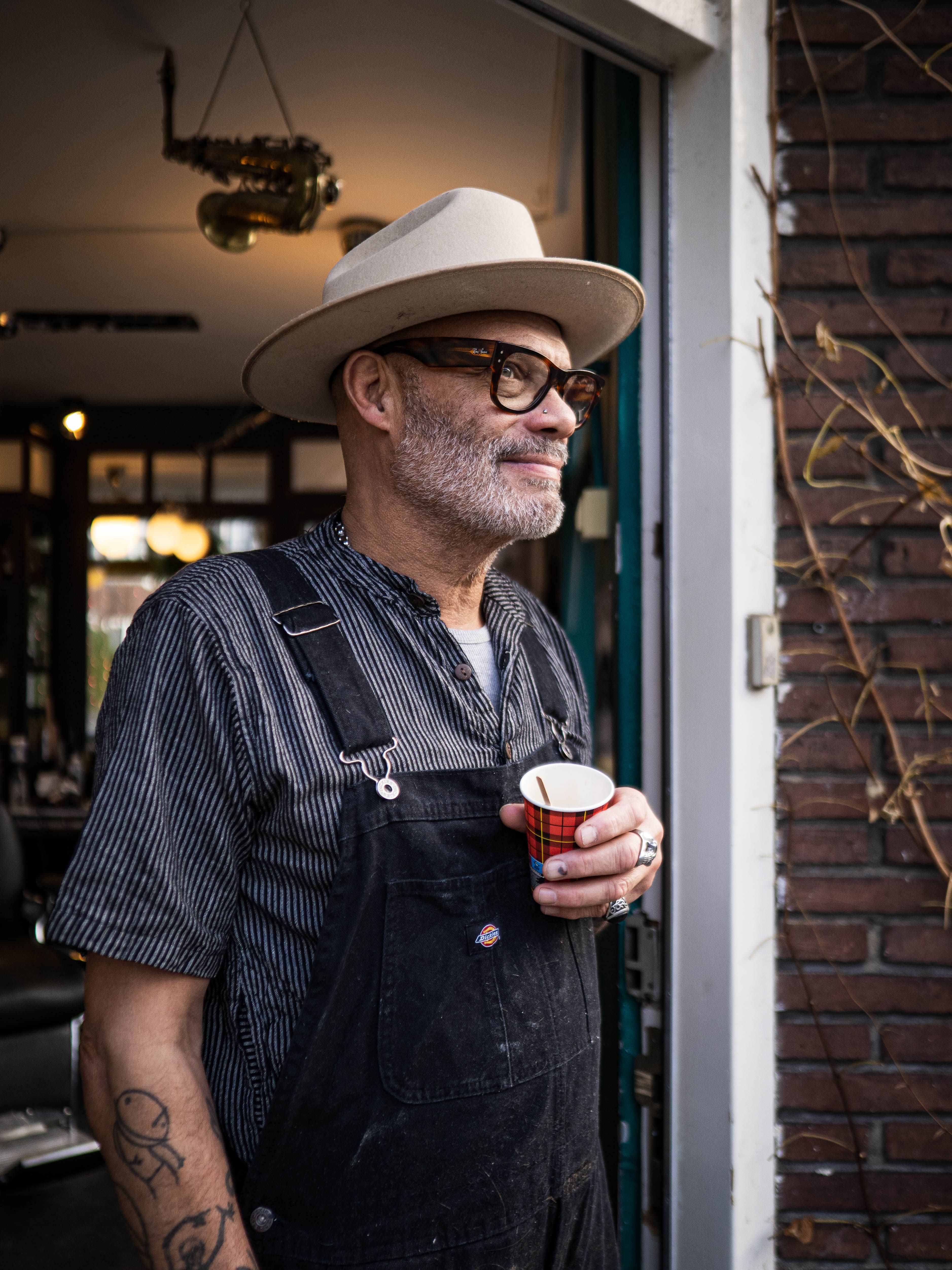 gratis Man staat in de deuropening met een kopje koffie, een hoed en een overall, in een stedelijke omgeving. Stockfoto