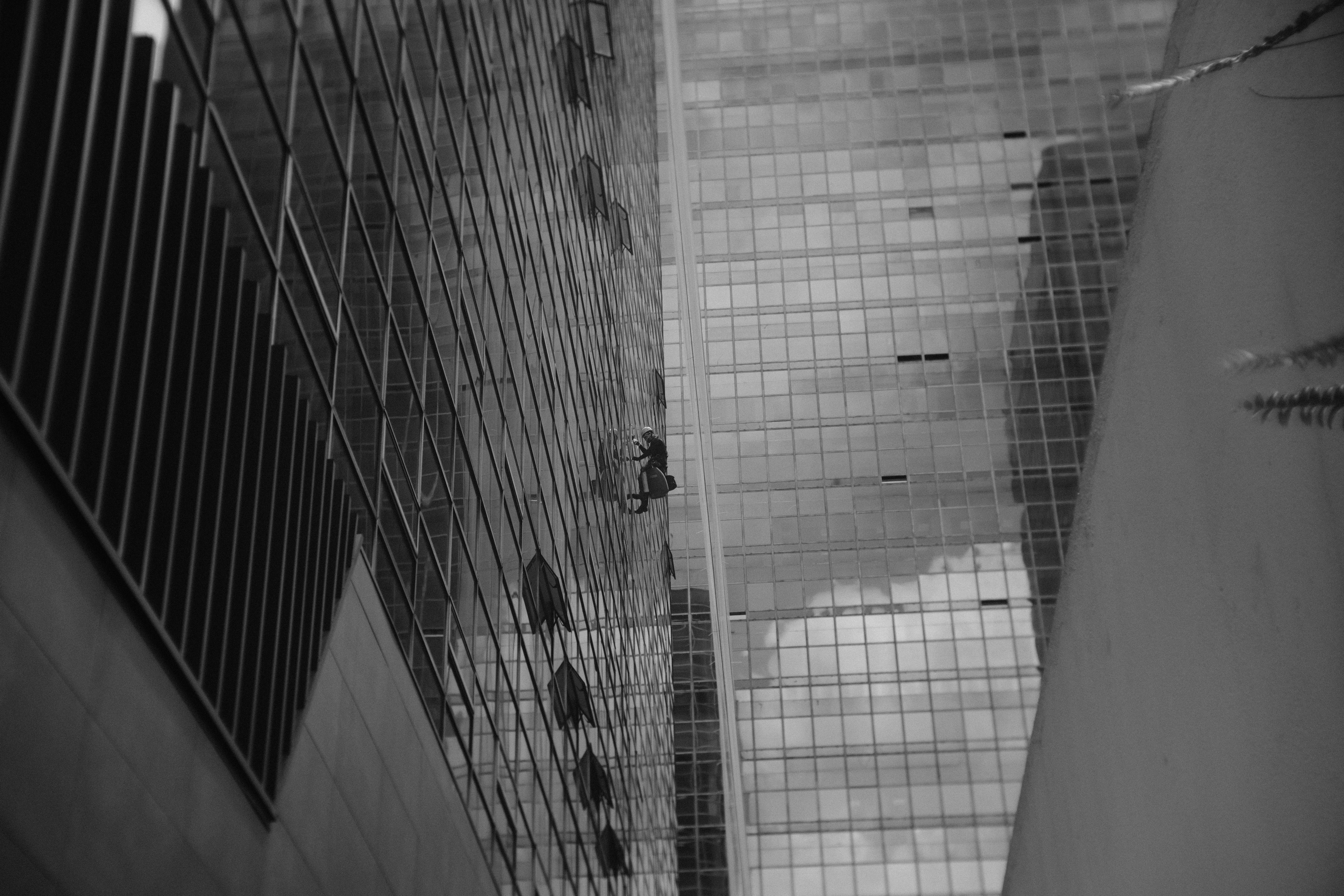 Bezpłatne Mycie okien przez okno. Osoba sprzątająca okna wspina się po ścianie wysokiego szklanego wieżowca, w otoczeniu miejskiej architektury. Zdjęcie z galerii