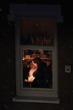 An adult studying in a warmly lit room, viewed through a window during a snowy night.