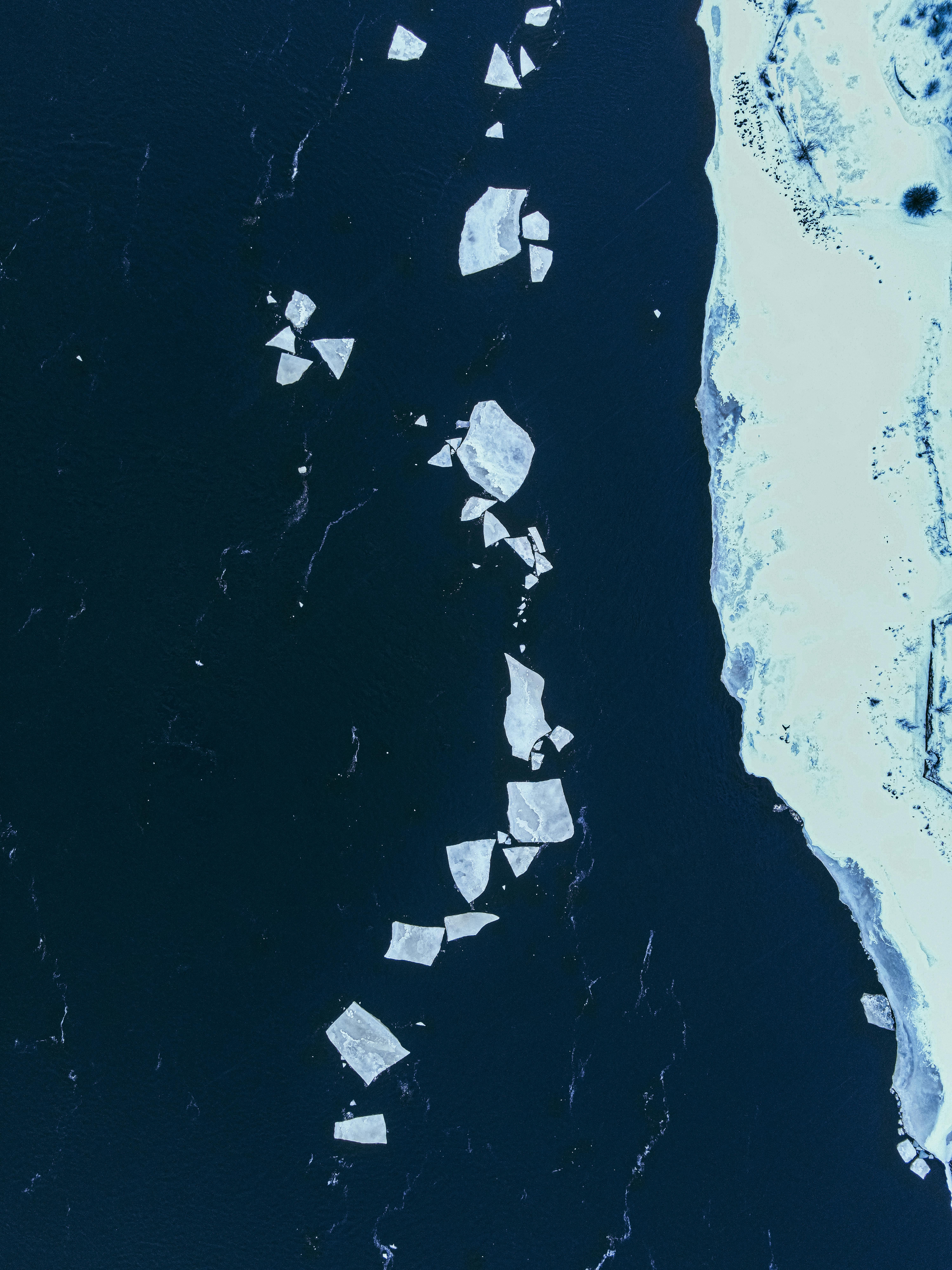 A stunning aerial shot of ice floes floating on dark water near a snowy shoreline.