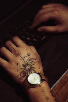 Close-up of tattooed hand with wristwatch typing on keyboard.