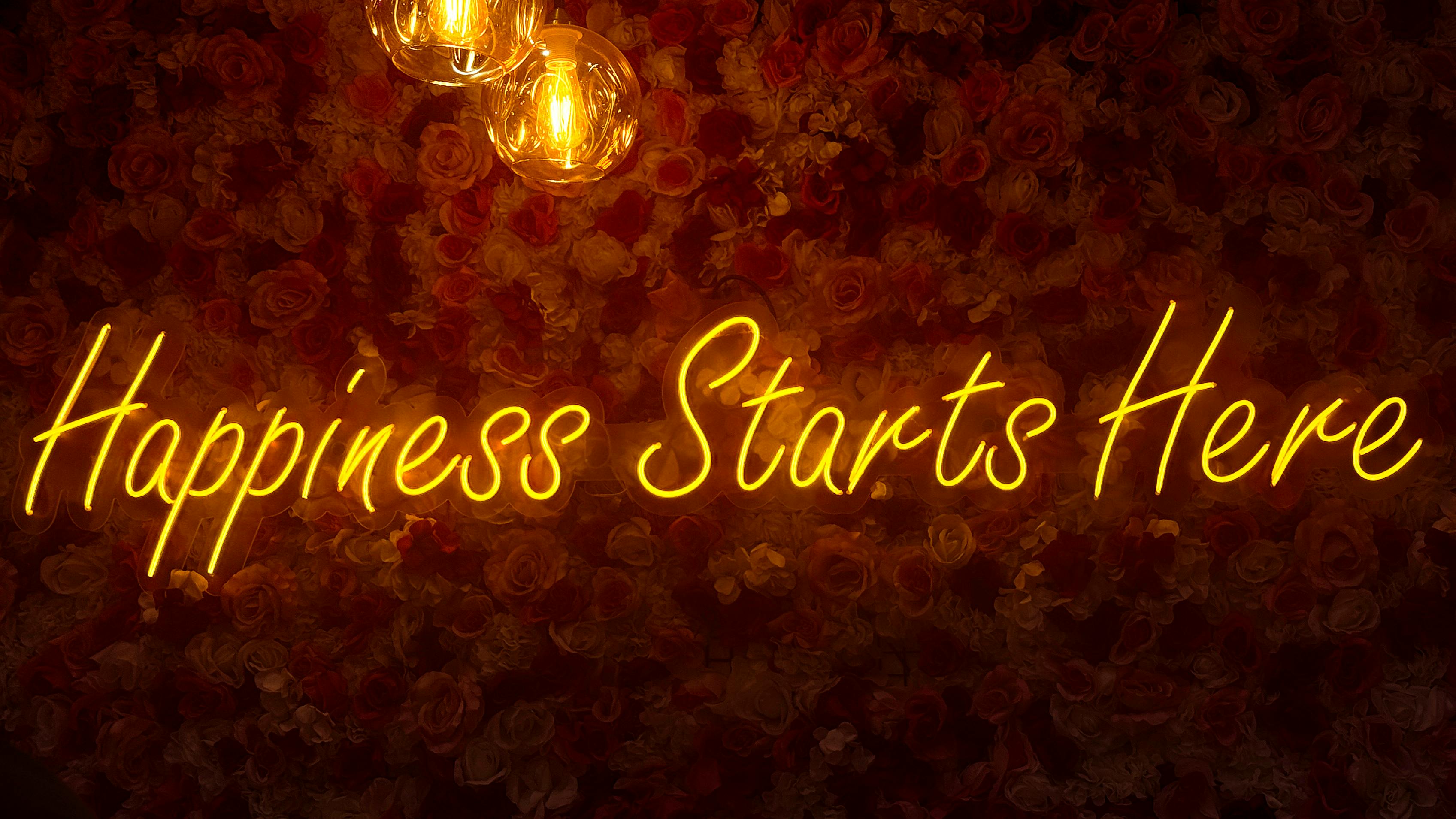 Neon sign 'Happiness Starts Here' illuminated on a floral wall with warm lighting.