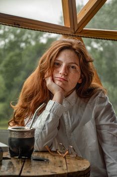 Redheaded woman relaxing indoors with mate drink on a wooden table, enjoying tranquility.