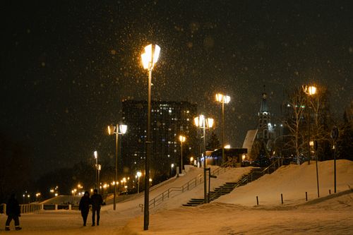 Free Snowfall in a city park at night with street lights and people walking, creating a serene winter atmosphere. Stock Photo