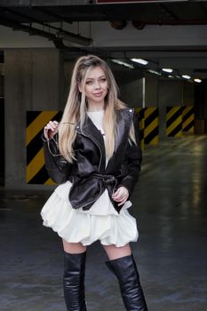 Stylish young woman poses in a parking garage wearing a leather jacket and boots.