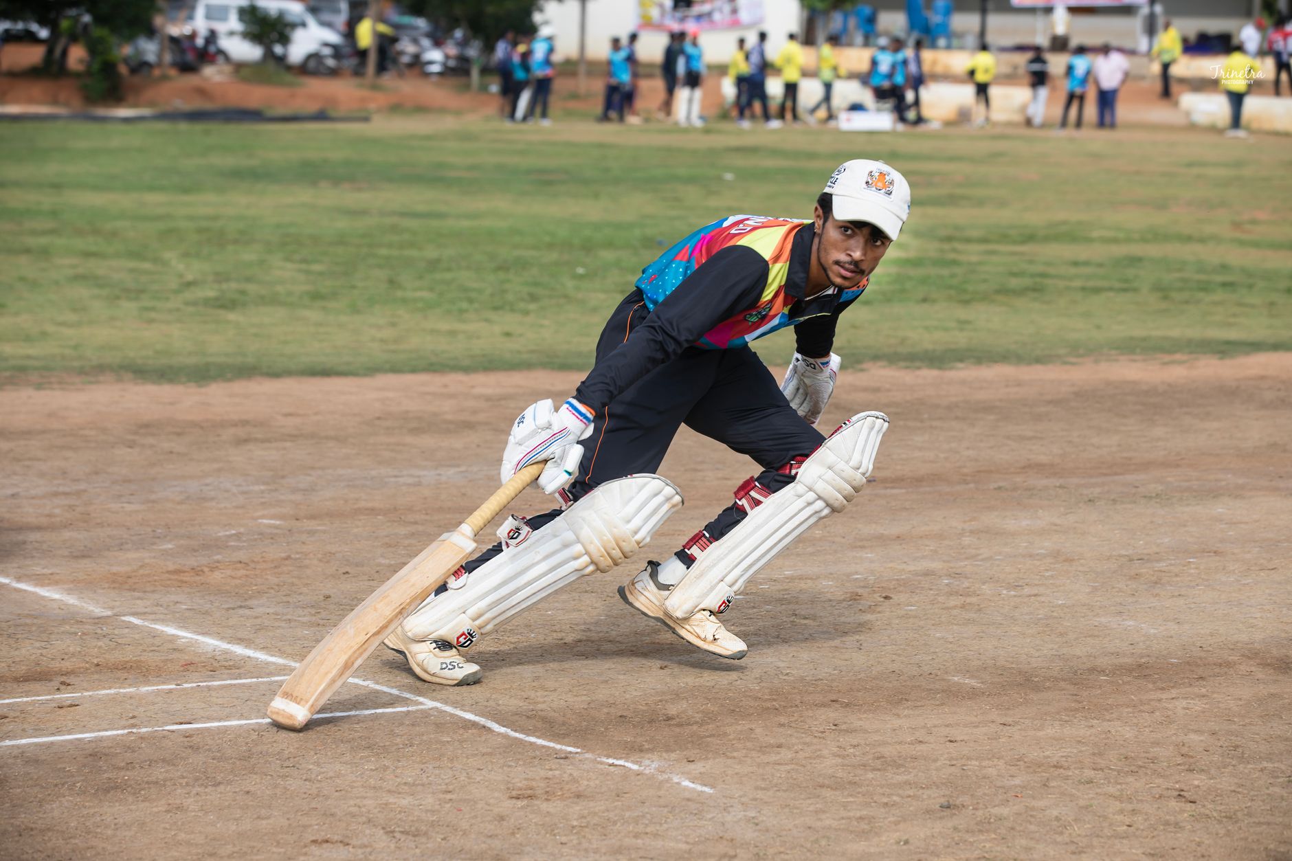 A cricket player in colorful attire bats during a daytime match on a sunny field.