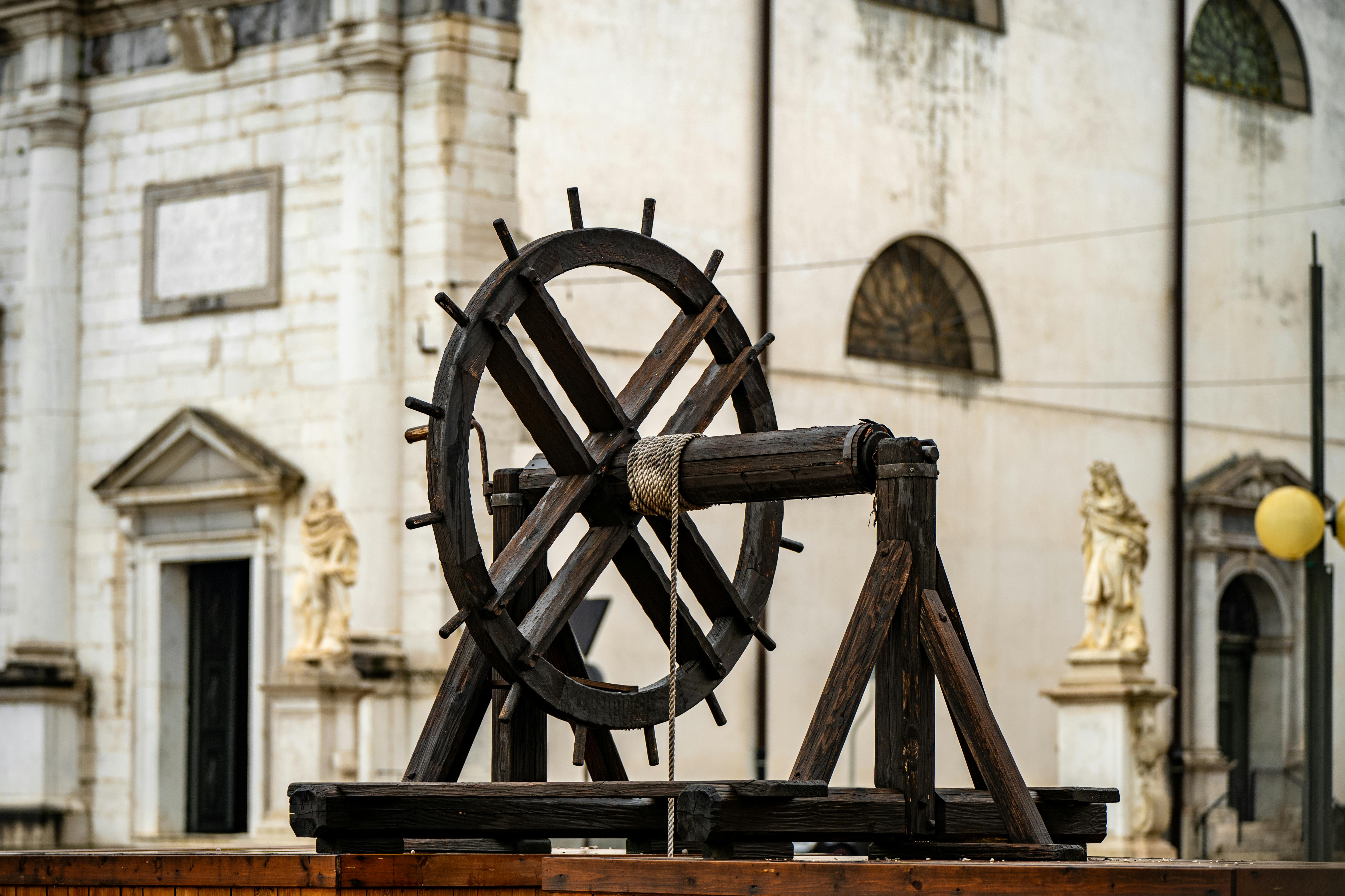 A historic wooden wheel in front of a classical European building with statues.