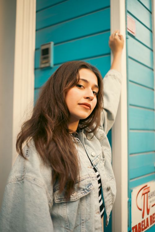 Free Young woman posing casually in front of a bright blue wall, wearing a denim jacket. Stock Photo