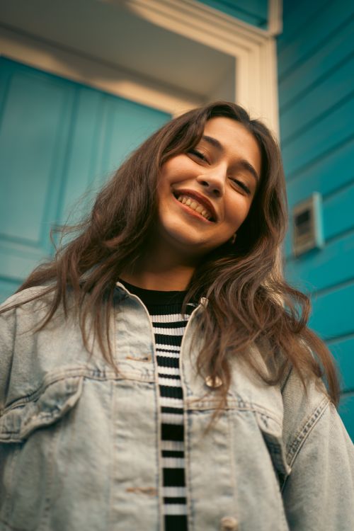 Free Portrait of a cheerful young woman in a denim jacket against a blue door, exuding positivity. Stock Photo
