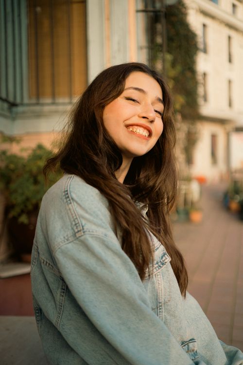 Free A young woman with a joyful smile wears a denim jacket while sitting outside in a warm setting. Stock Photo