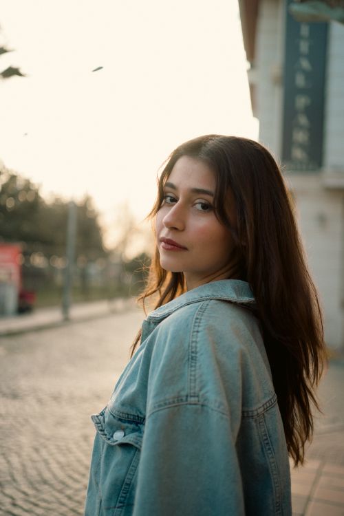 Free A woman in a denim jacket strolls down a cobblestone street at sunset. Stock Photo