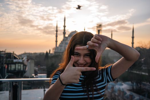 Smiling woman frames view of Istanbul's Blue Mosque at sunset, capturing joyful travel moment.