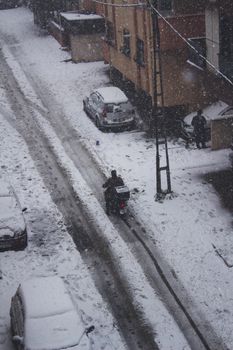A winter scene of a motorcyclist navigating a snowy street in an urban neighborhood.