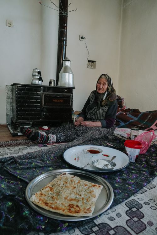 Free Elderly woman sitting near a stove enjoying a traditional meal in a cozy indoor setting. Stock Photo