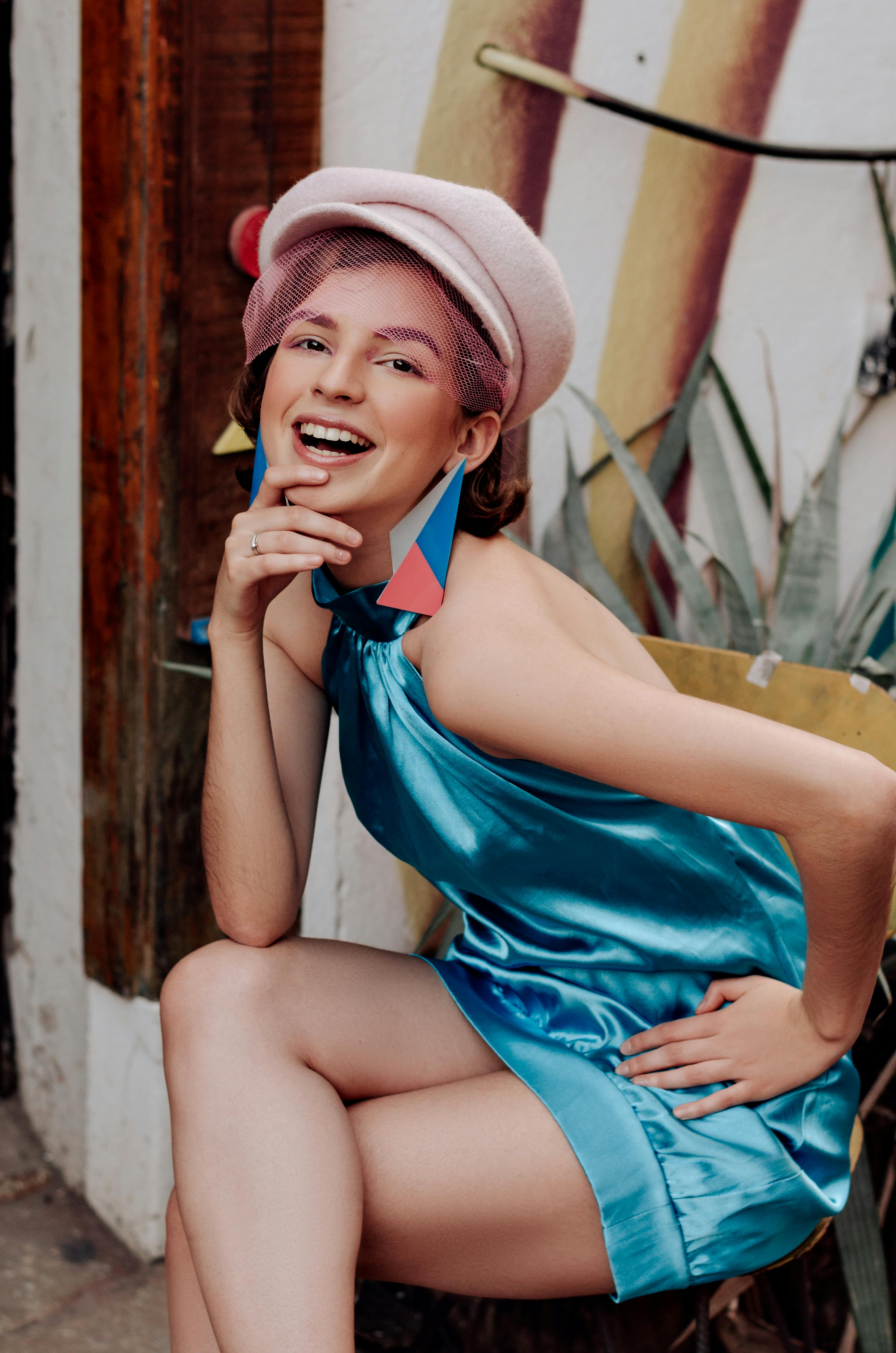 一位时尚的年轻女子身着活力蓝色连衣裙，头戴别致帽子，在户外微笑着摆好姿势拍照