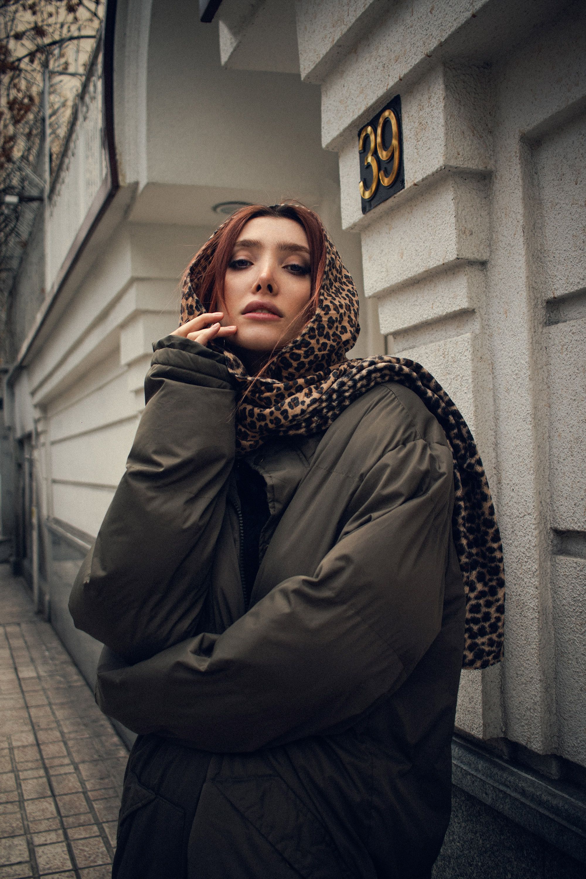 Fashionable woman with leopard print scarf poses outdoors by a numbered building.