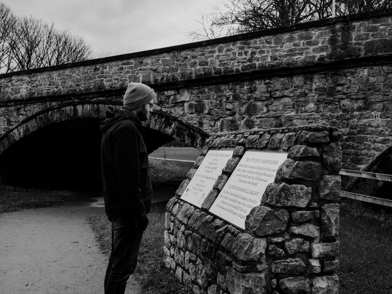 A man stands reading informational plaques by an old stone bridge - historical marker database