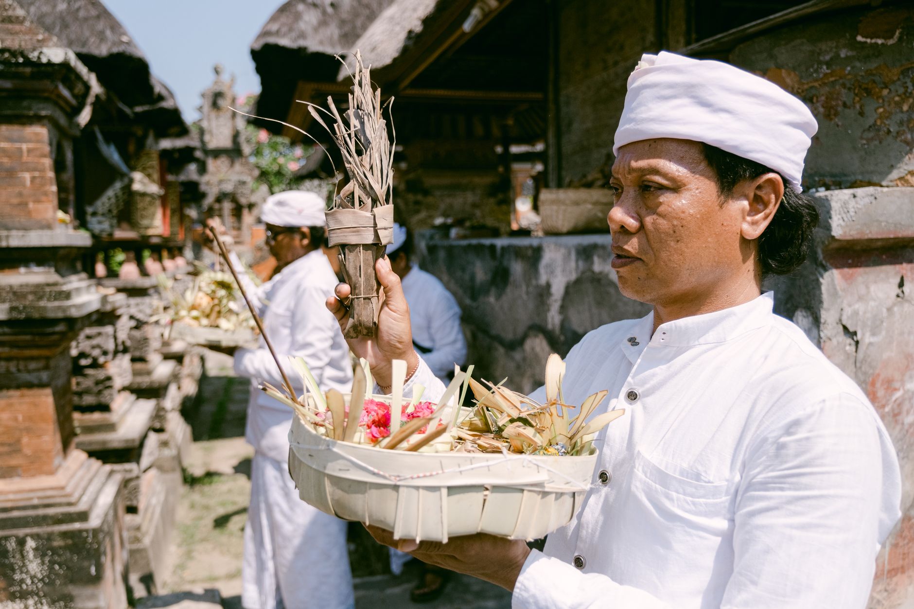 Balinese priest in white holding traditional offerings during a ceremony in a temple courtyard.