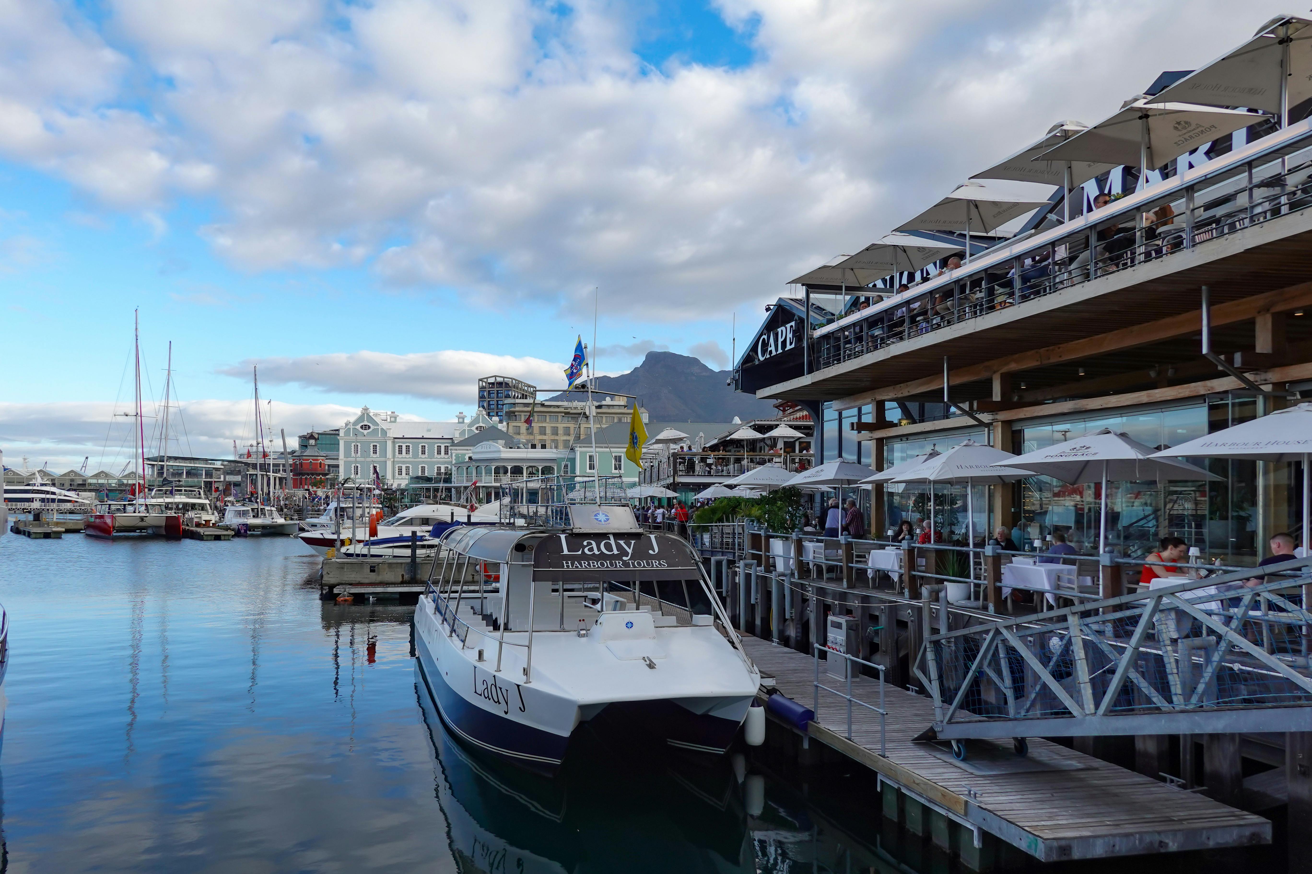 Explore the bustling harbor scene of Cape Town with docked boats and waterfront dining under a clear sky.