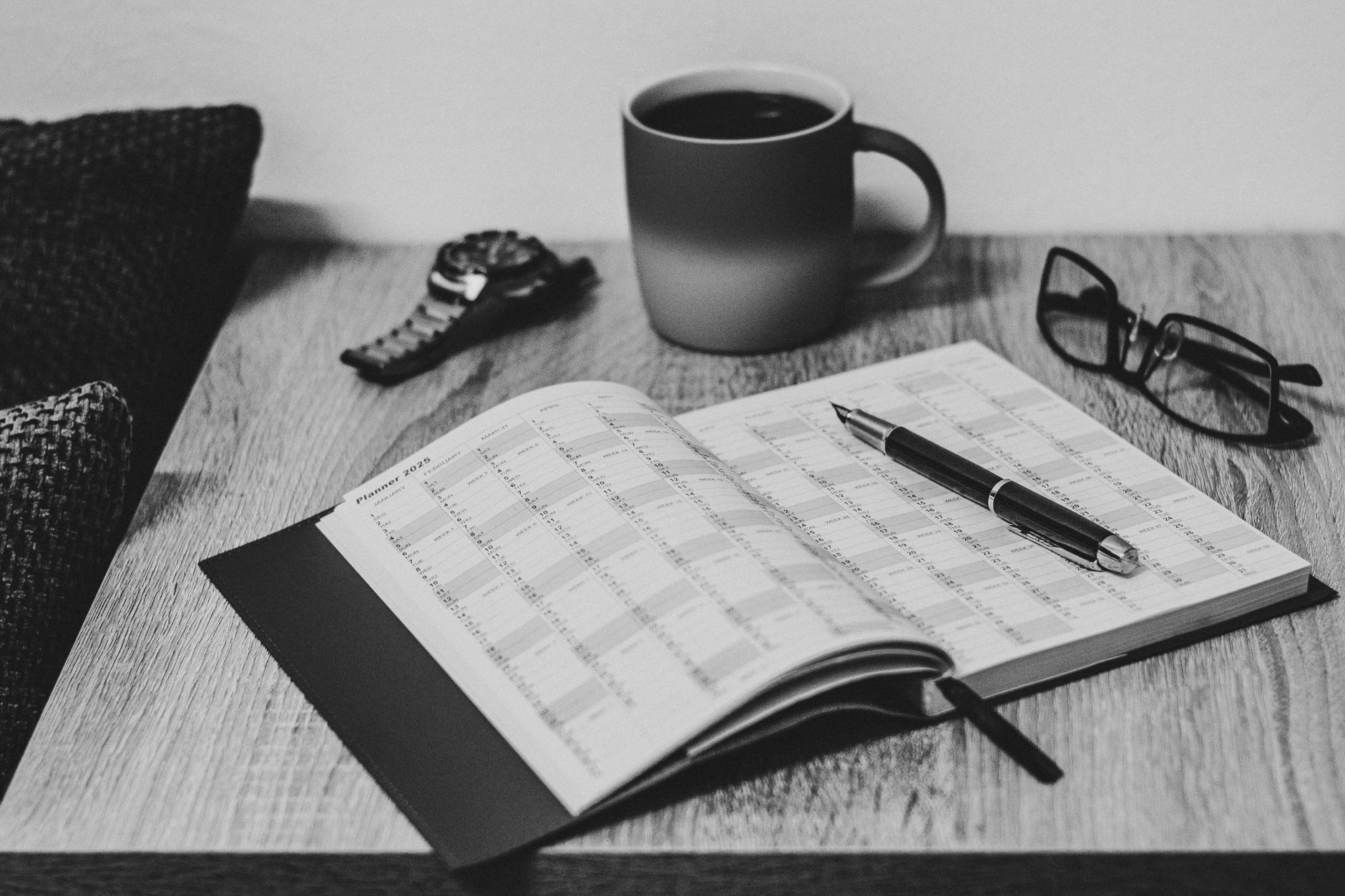 Monochrome image of a desk featuring an open planner, pen, coffee cup, and glasses.