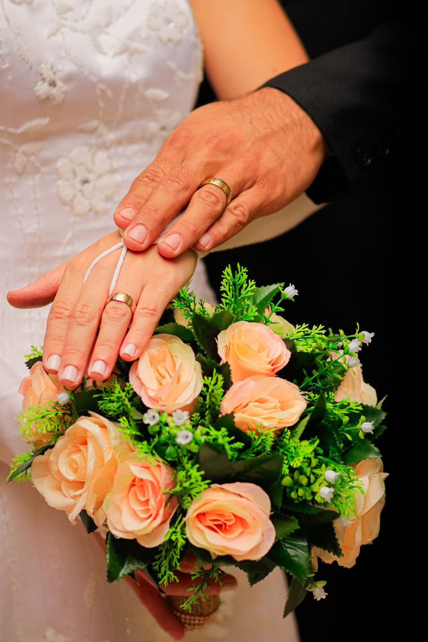 Close-up of a bride and groom's hands showcasing wedding rings with a lush bouquet.