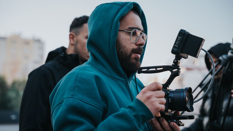 Filmmaker in blue hoodie operating a camera outdoors with a screen monitor attached.