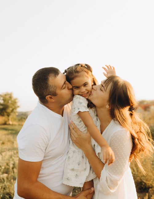 Free Loving parents embrace their cheerful daughter in a sunny outdoor setting, capturing a warm family moment. Stock Photo