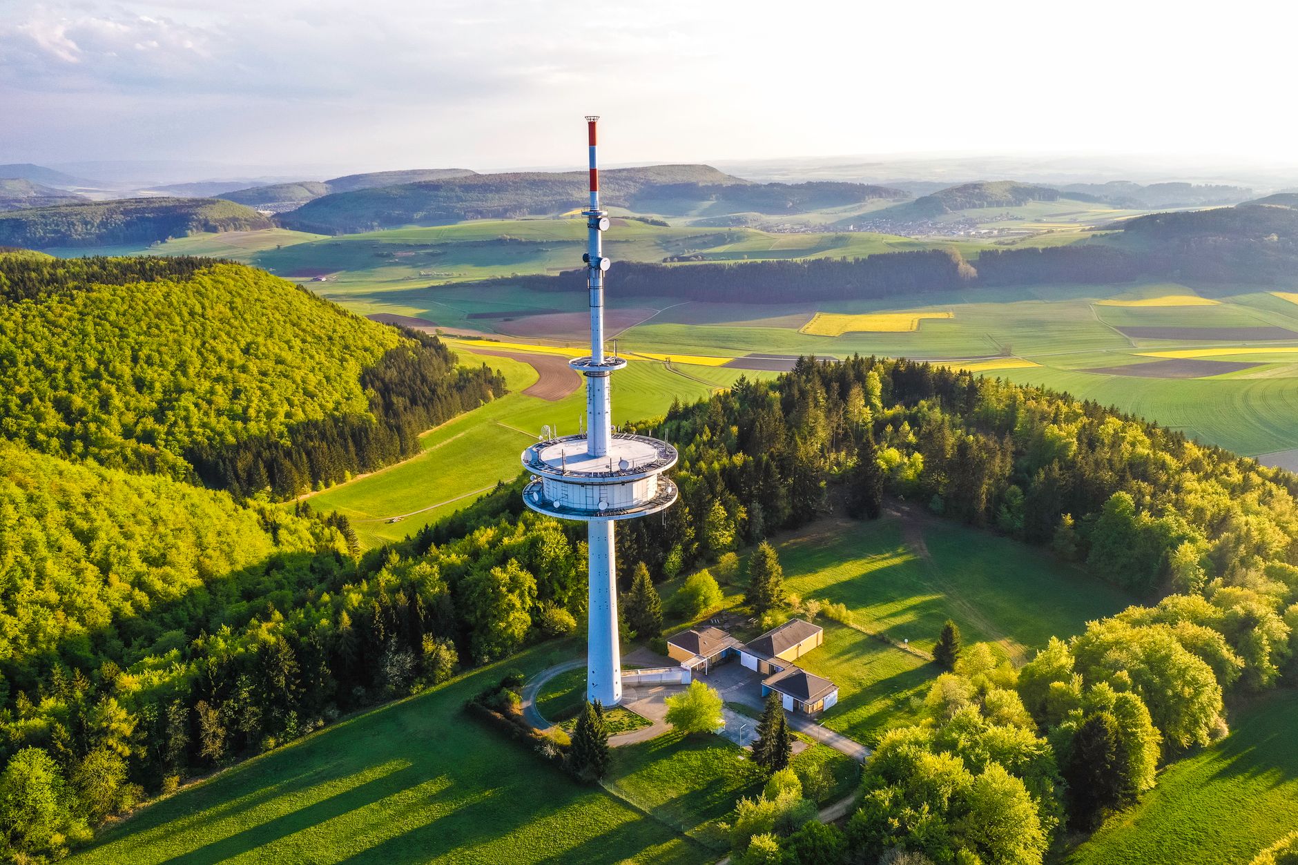 Aerial shot of a communication tower amidst lush greenery and open fields during daytime.