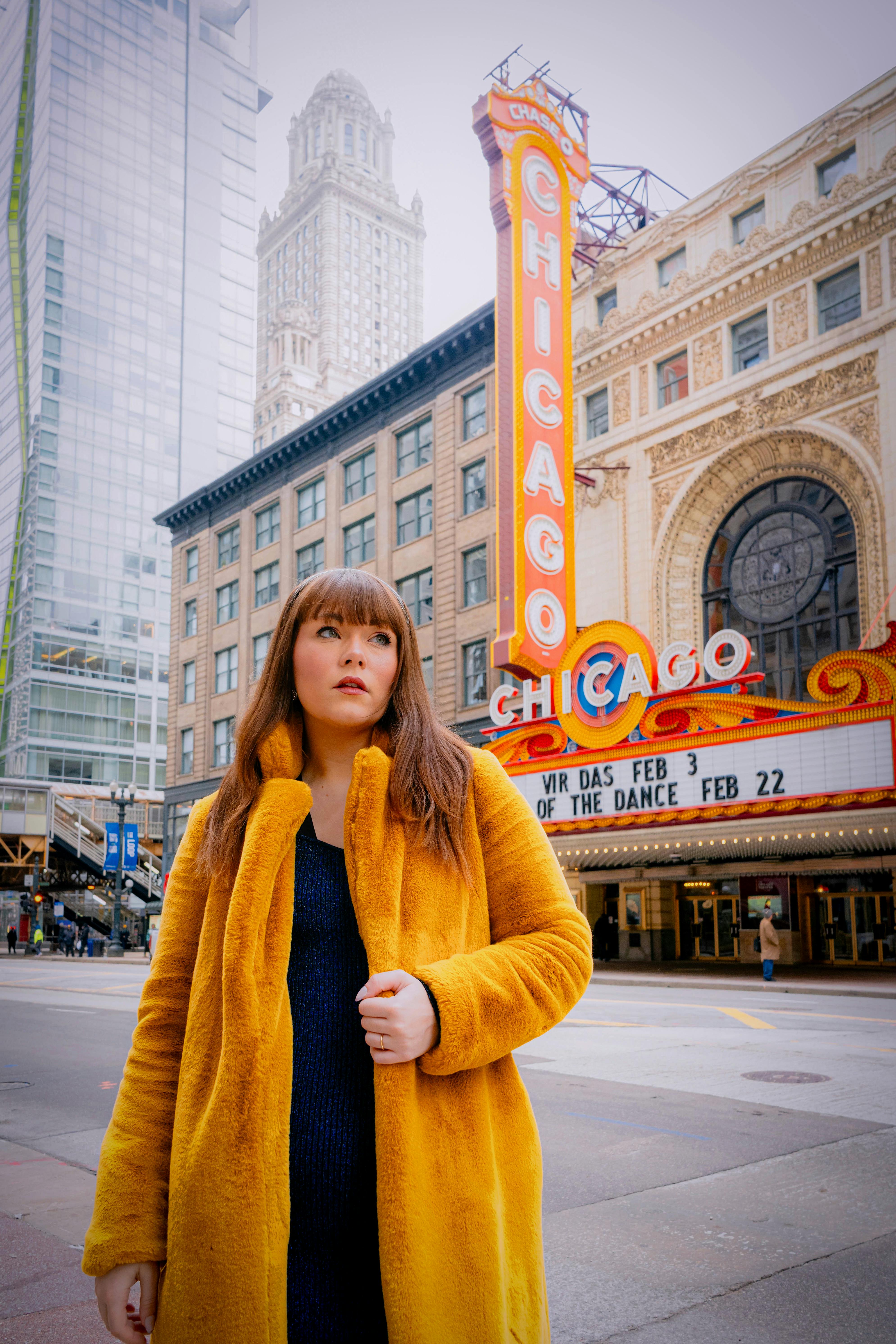 Young woman in a yellow coat standing in front of Chicago Theater marquee on a city street.