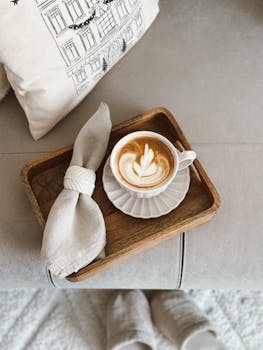Cozy latte in wooden tray with napkin and pillow background, perfect for relaxing moments.