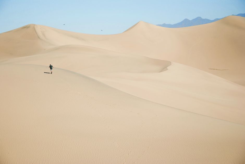 Solitary figure traversing expansive sand dunes under a clear blue sky, showcasing vast desert landscapes.
