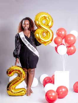 A woman celebrates her 28th birthday with balloons and a sash indoors.