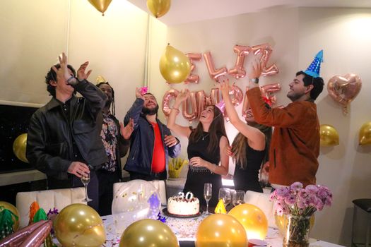 Group of friends celebrating a birthday indoors, surrounded by balloons and cake.