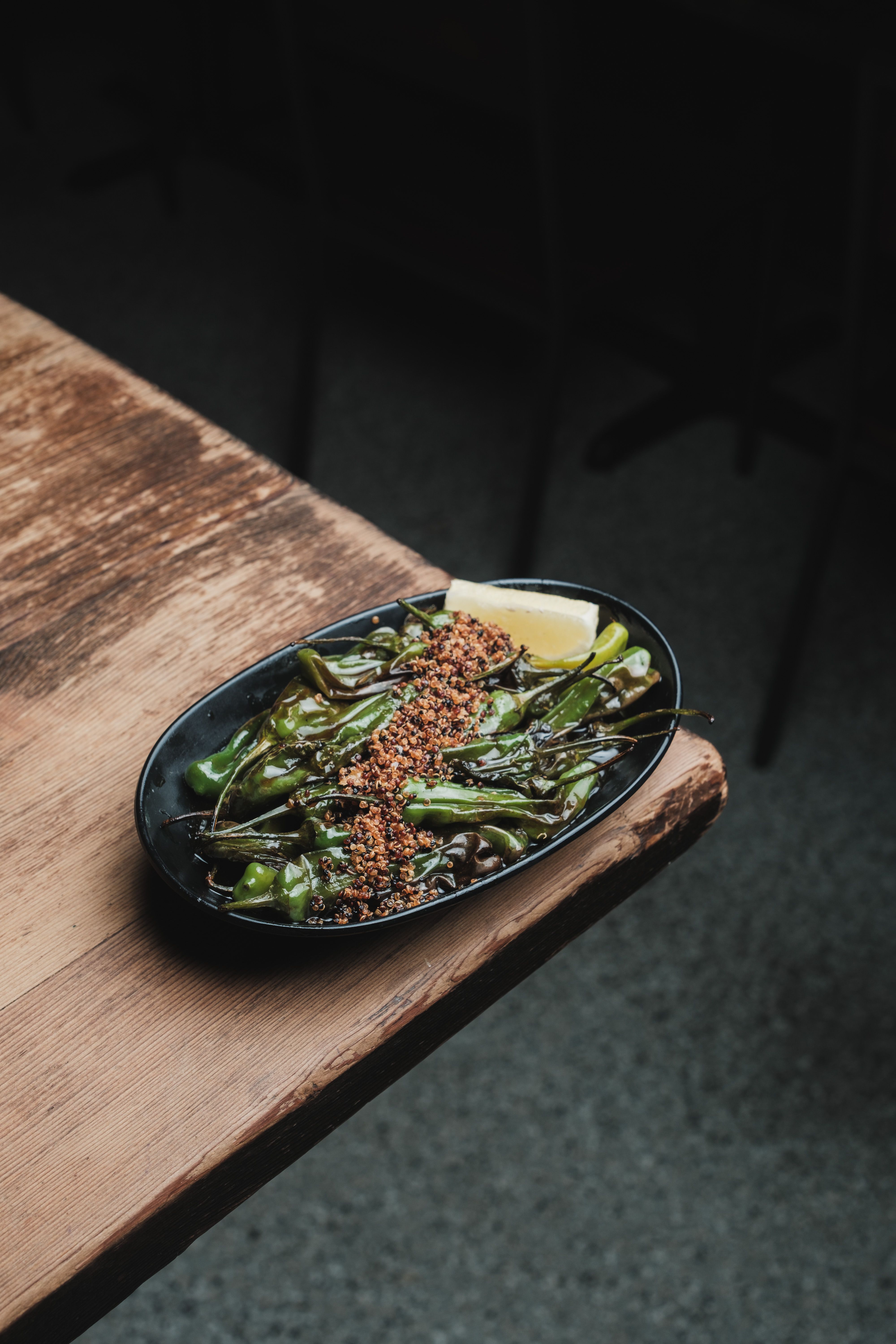 A pile of blistered, charred shishito peppers on a white plate, sprinkled with flaky salt and a wedge of lemon on the side.