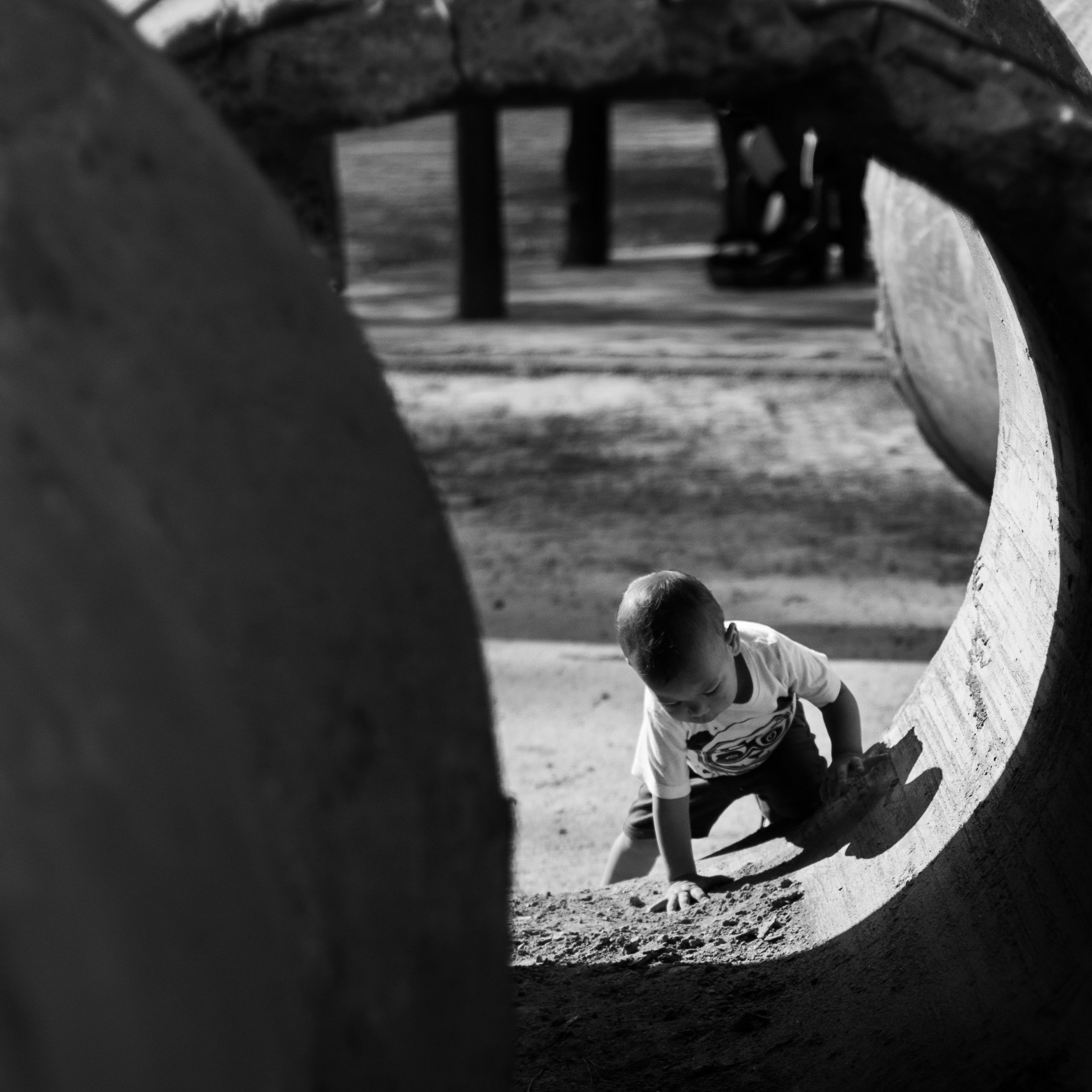 Black and white photo of a child crawling through a large concrete tube at a playground.