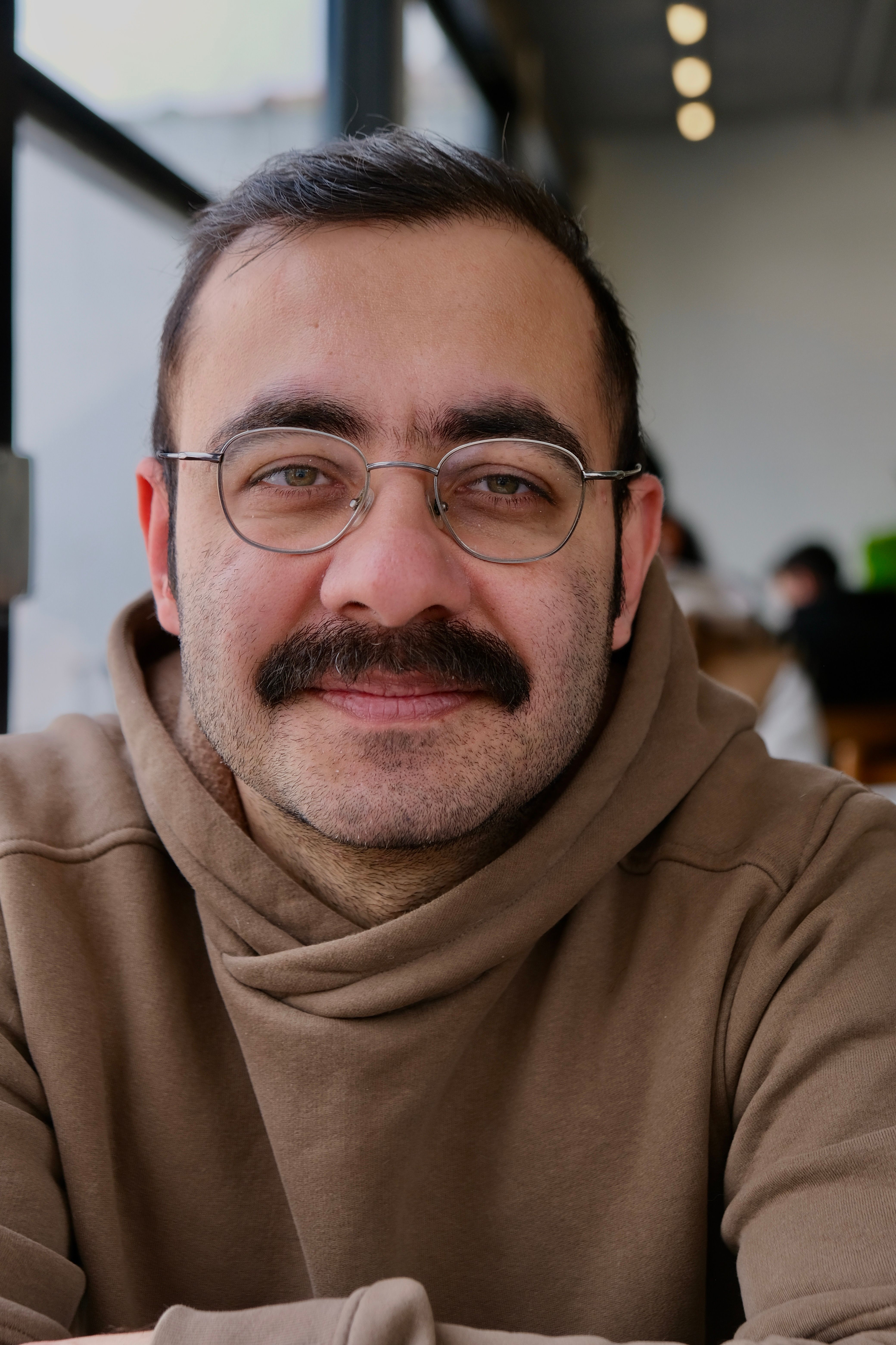 Close-up portrait of a smiling man with glasses and mustache in a cozy Istanbul cafe.